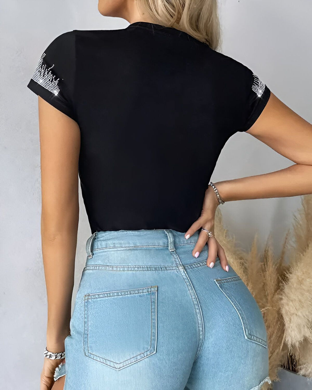 Une femme porte un t-shirt noir moulant à manches courtes avec des strass sur les épaules et un jean bleu clair taille haute.