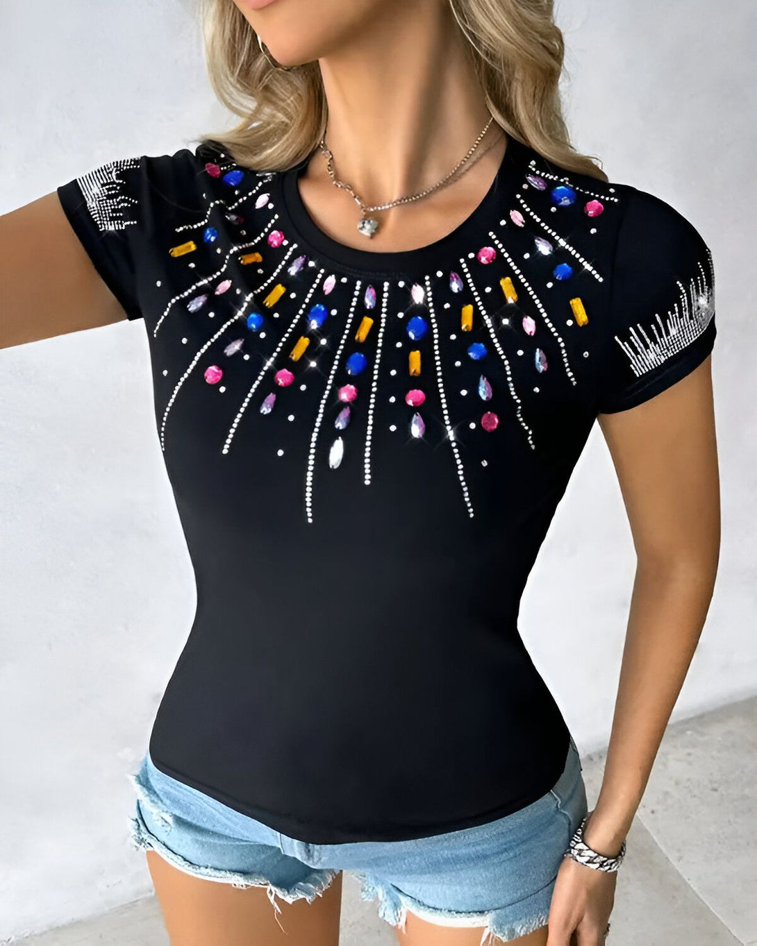Une femme porte un t-shirt noir avec des pierres précieuses colorées sur le devant. Le t-shirt est moulant et a des manches courtes. La femme est blonde et porte un collier avec un pendentif en forme de cœur. Elle est également maquillée et a les ongles manucurés.