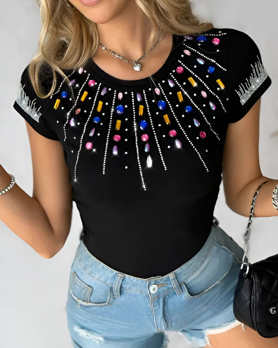 Une femme porte un t-shirt noir avec des strass multicolores. Le t-shirt est moulant et a des manches courtes. La femme est blonde et porte un collier avec un pendentif en forme de cœur. Elle porte également un jean bleu déchiré et des talons hauts noirs.
