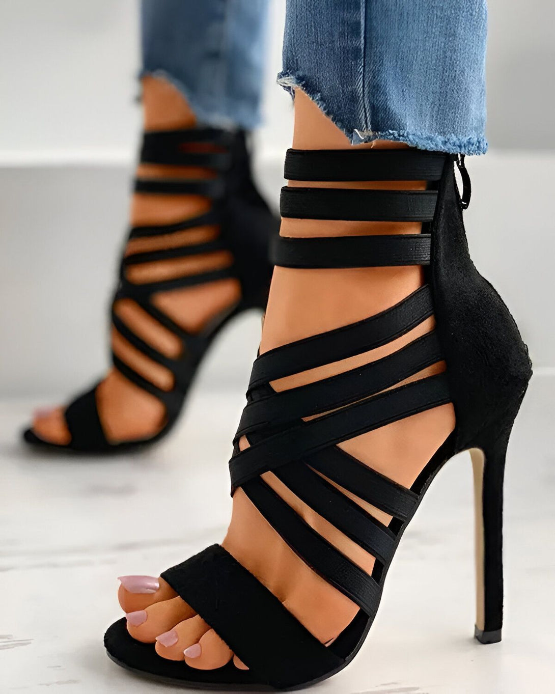 Une femme porte des sandales à talons hauts noires. Les sandales sont attachées à la cheville et ont des brides qui s'enroulent autour de la jambe. Le talon est fin et mesure environ 10 centimètres. La femme porte un jean bleu et les ongles des pieds peints en rose.