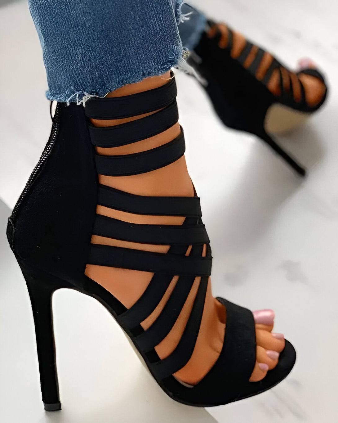 Une femme porte des sandales à talons hauts noires. Les sandales sont en daim et ont des brides qui s'enroulent autour de la cheville. La femme porte un jean bleu et les ongles des pieds peints en rose.