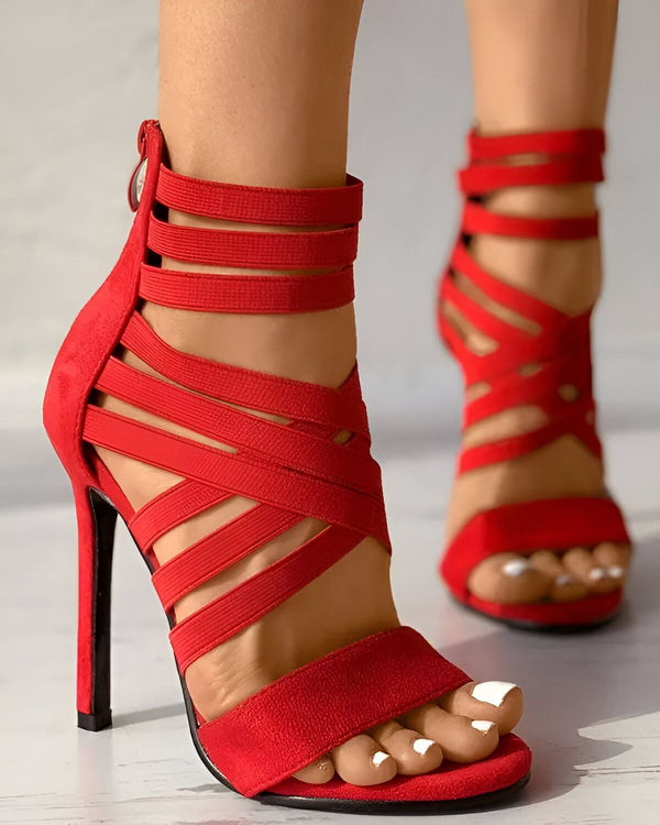 Une femme porte des sandales rouges à talons hauts. Les sandales sont en daim et ont des brides qui s'enroulent autour de la cheville et du pied. Le talon est fin et mesure environ 10 centimètres. La femme a les ongles des pieds peints en blanc.