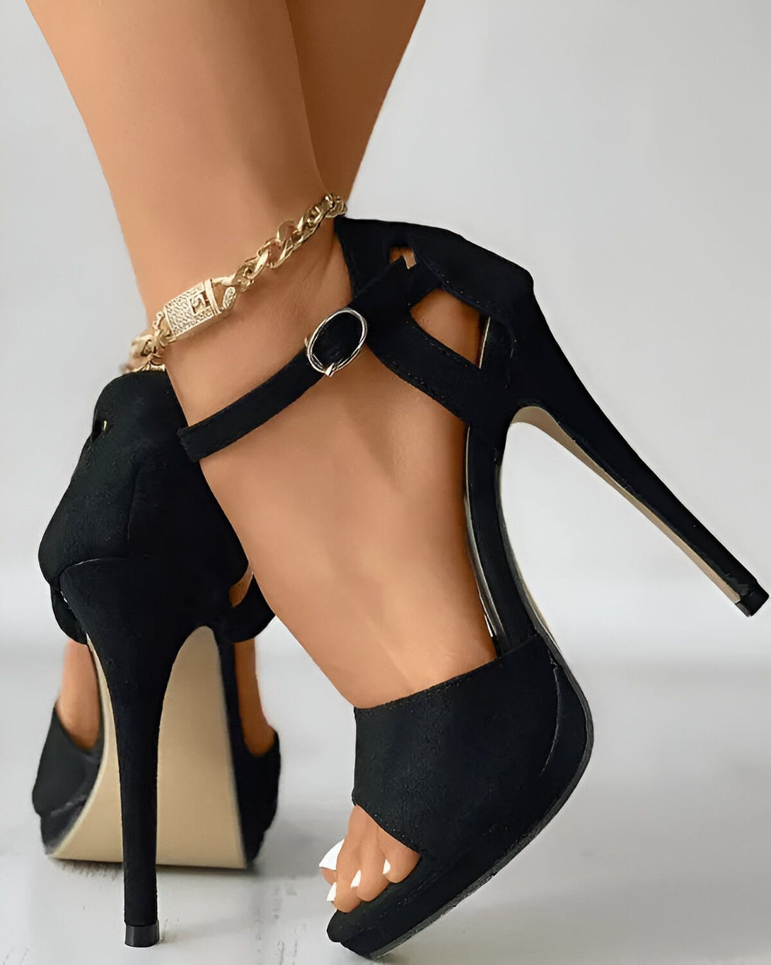 Une femme porte des sandales à talons hauts noires. Les sandales sont en daim et ont une bride à la cheville. La femme a les ongles des pieds peints en blanc.