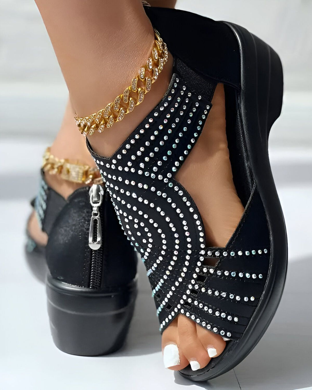 Une femme porte des sandales noires à talons compensés. Les sandales sont ornées de strass et ont une fermeture éclair sur le côté. La femme porte également une chaîne de cheville en or.