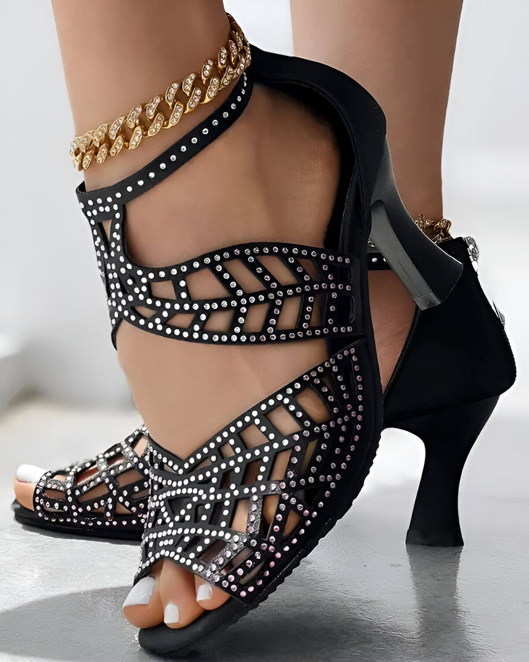 Une image de chaussures à talons noirs avec des strass roses et argentés. Les chaussures sont portées par une femme avec une chaîne dorée à la cheville.