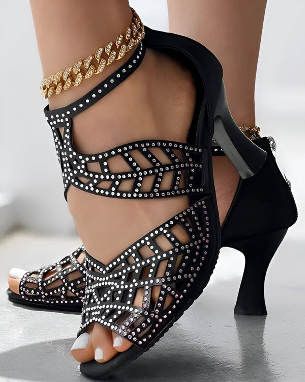 Une image de chaussures à talons noirs avec des strass roses et argentés. Les chaussures sont portées par une femme avec une chaîne dorée à la cheville.