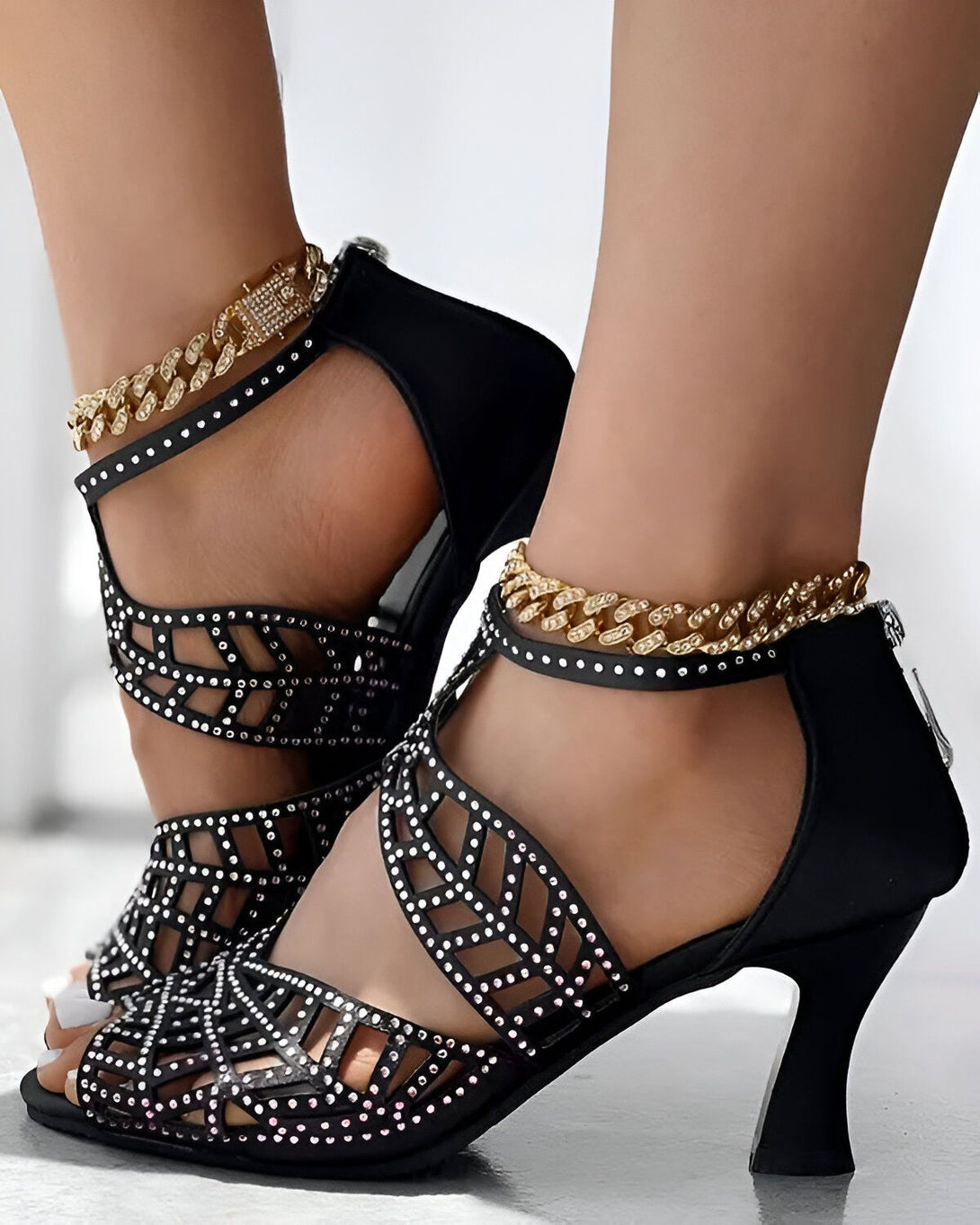 Une femme porte des sandales noires à talons hauts. Les sandales sont ornées de strass et de chaînes dorées.