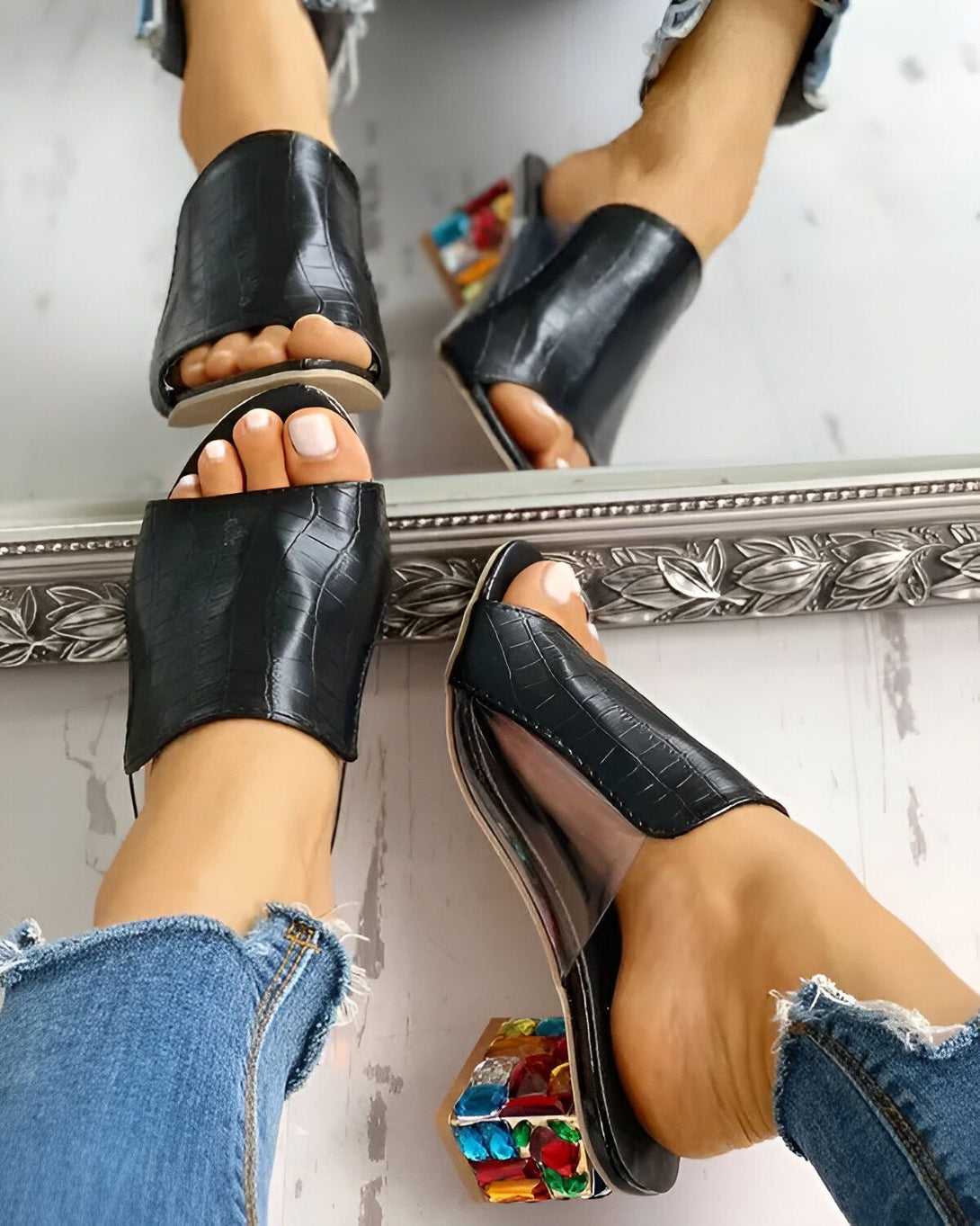 Une femme porte des mules noires à talons hauts transparents. Les talons sont décorés de pierres précieuses multicolores. La femme est assise sur le bord d'un lit et porte un jean bleu.