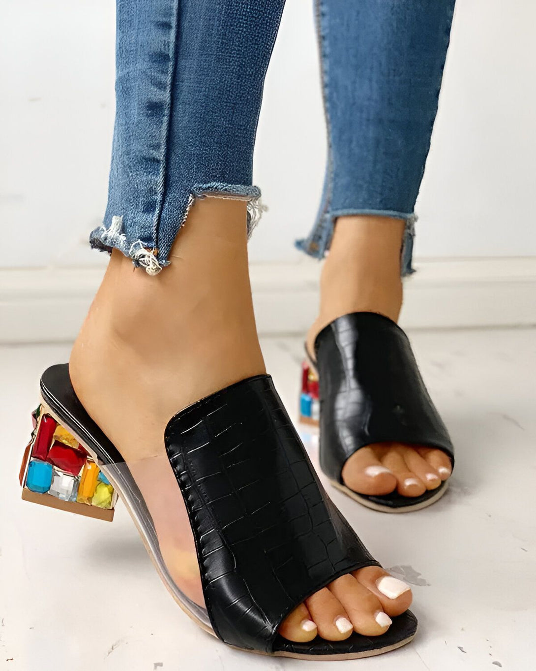 Une femme porte des mules noires à talons transparents. Les talons sont décorés de pierres précieuses multicolores. La femme porte un jean bleu déchiré.