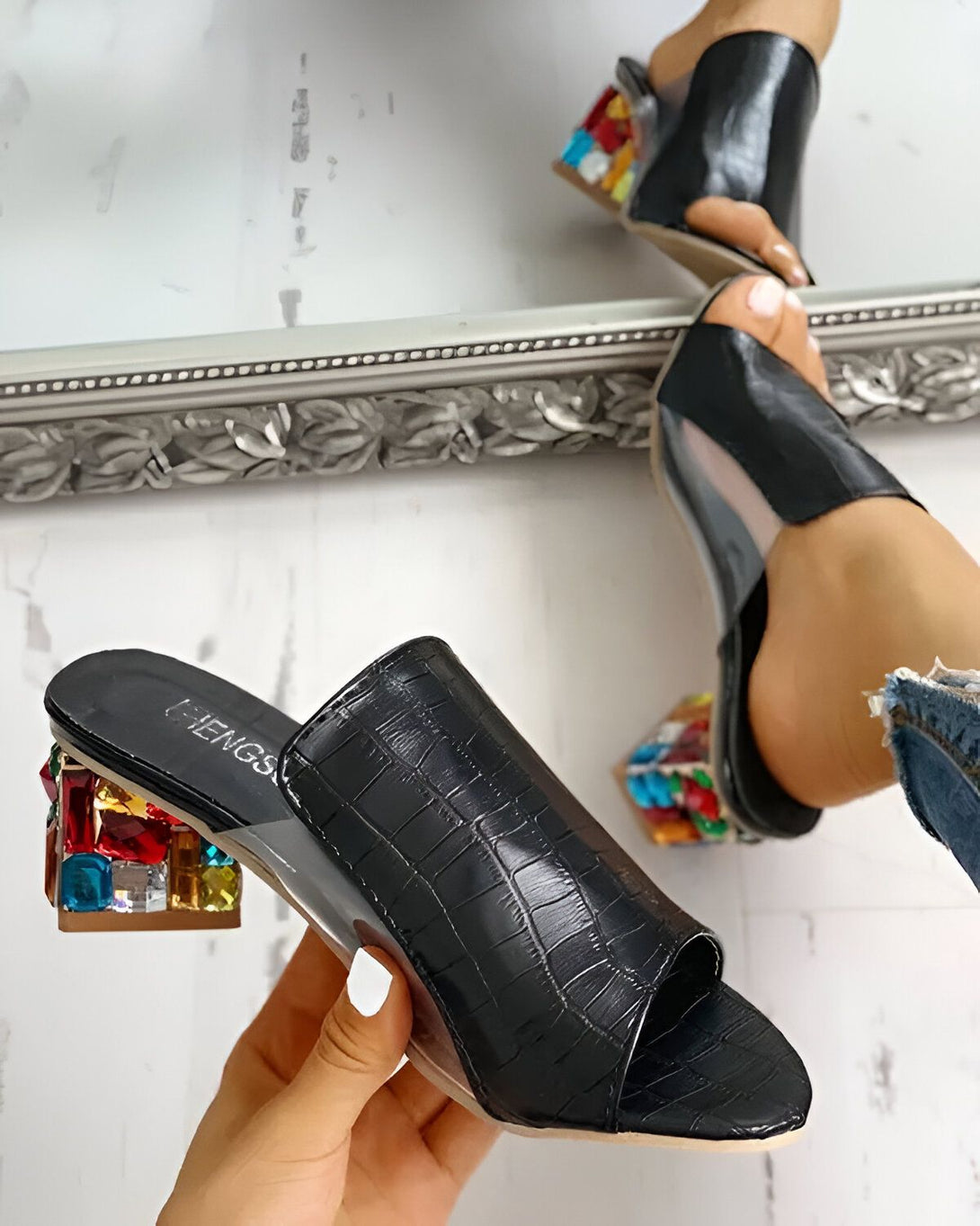 Une image d'une paire de mules noires à talons hauts avec des pierres précieuses colorées sur le talon. Les mules sont portées par une femme qui porte des jeans bleus. L'arrière-plan est un miroir.