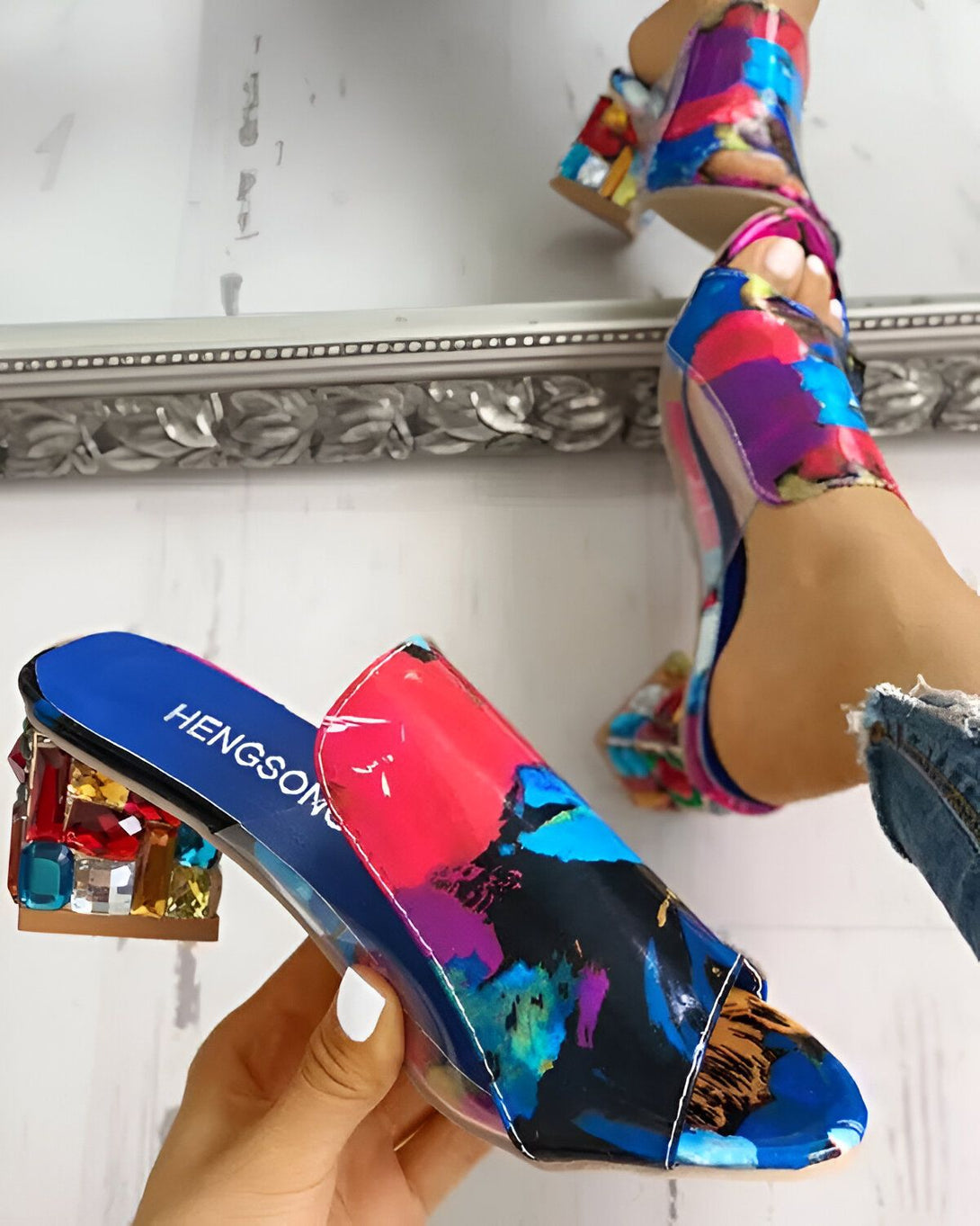 Une femme porte des mules à talons hauts multicolores. Les mules sont transparentes et ont un talon en forme de bloc. La femme porte un jean bleu et un haut rose. Elle est debout devant un miroir.