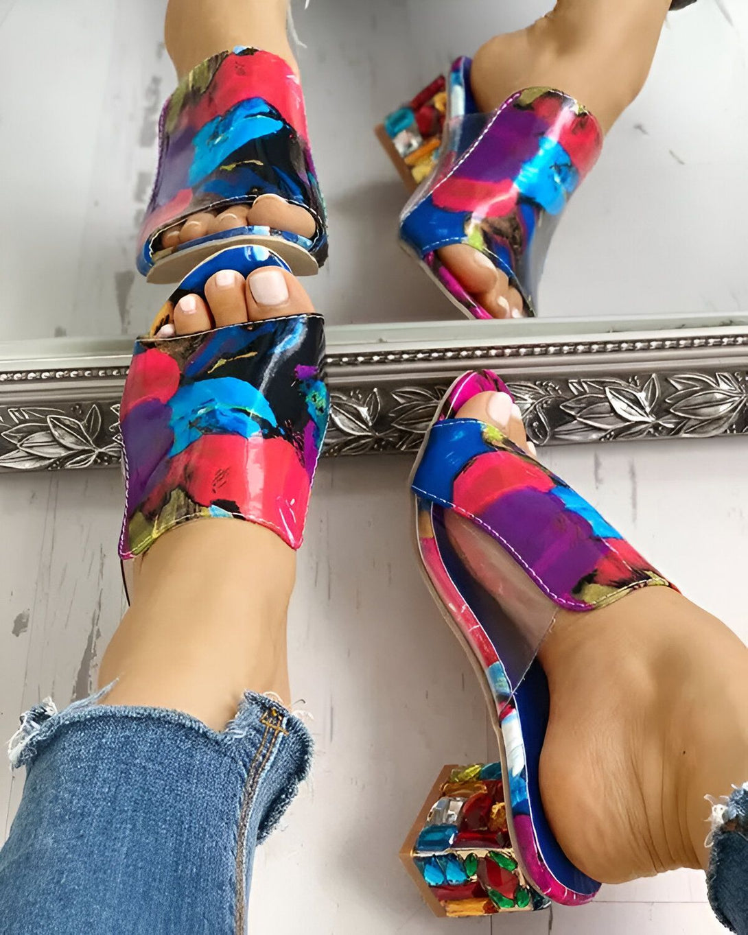 Une femme porte des mules à talons hauts transparentes avec un motif floral coloré. Les mules ont une bride transparente et un talon en forme de bloc avec des détails en strass. La femme porte un jean bleu déchiré et les ongles des pieds peints en blanc. Elle est debout devant un miroir et l'on peut voir son reflet dans le miroir.