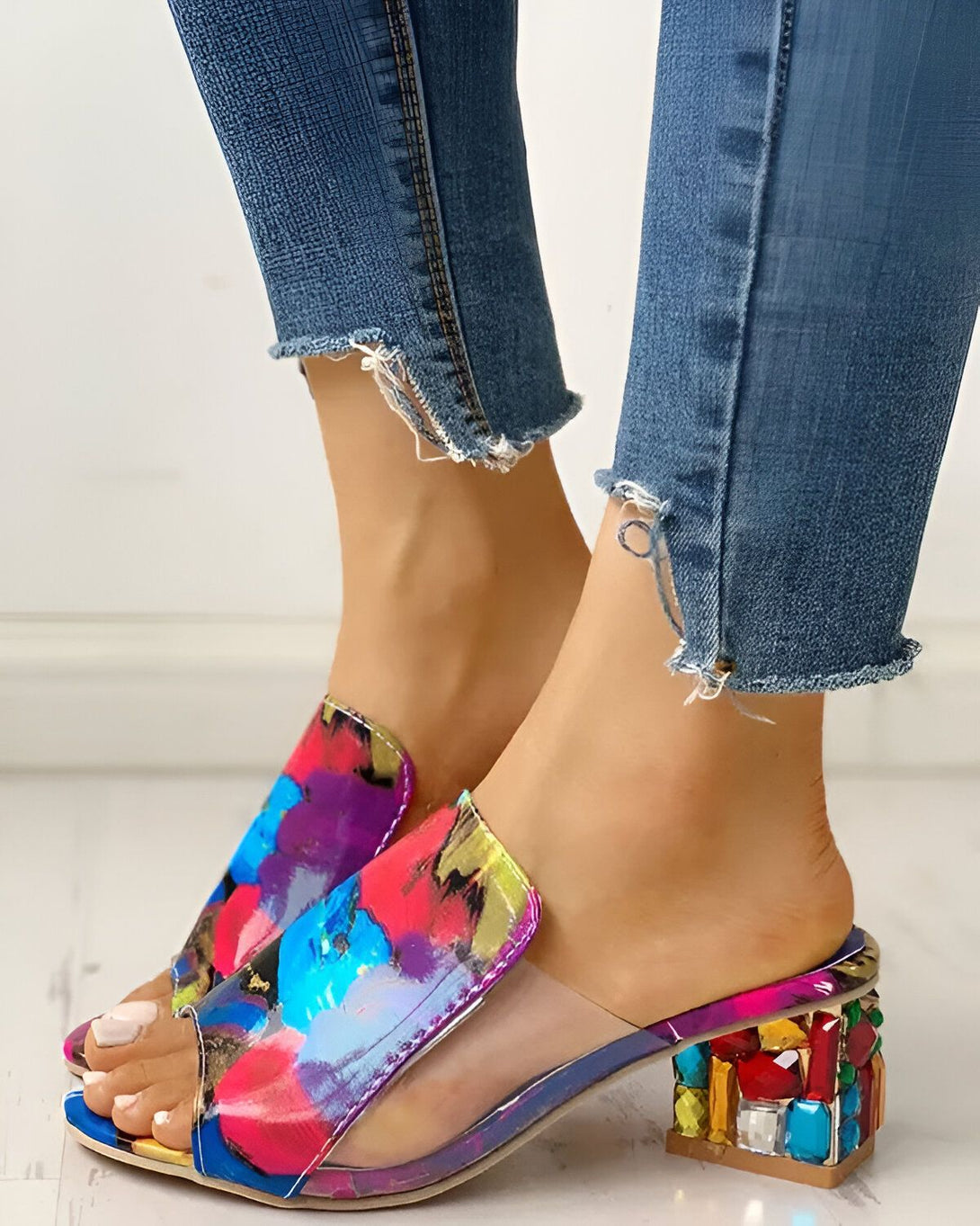 Une femme porte des mules à talons hauts transparentes avec un motif coloré et des pierres précieuses sur le talon. Elle porte un jean bleu troué.