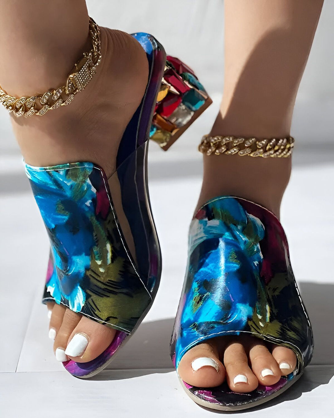 Une femme porte des mules à talons hauts bleues et blanches à fleurs. Les mules sont ornées de strass multicolores. La femme a les ongles des pieds blancs et porte des bracelets de cheville dorés.