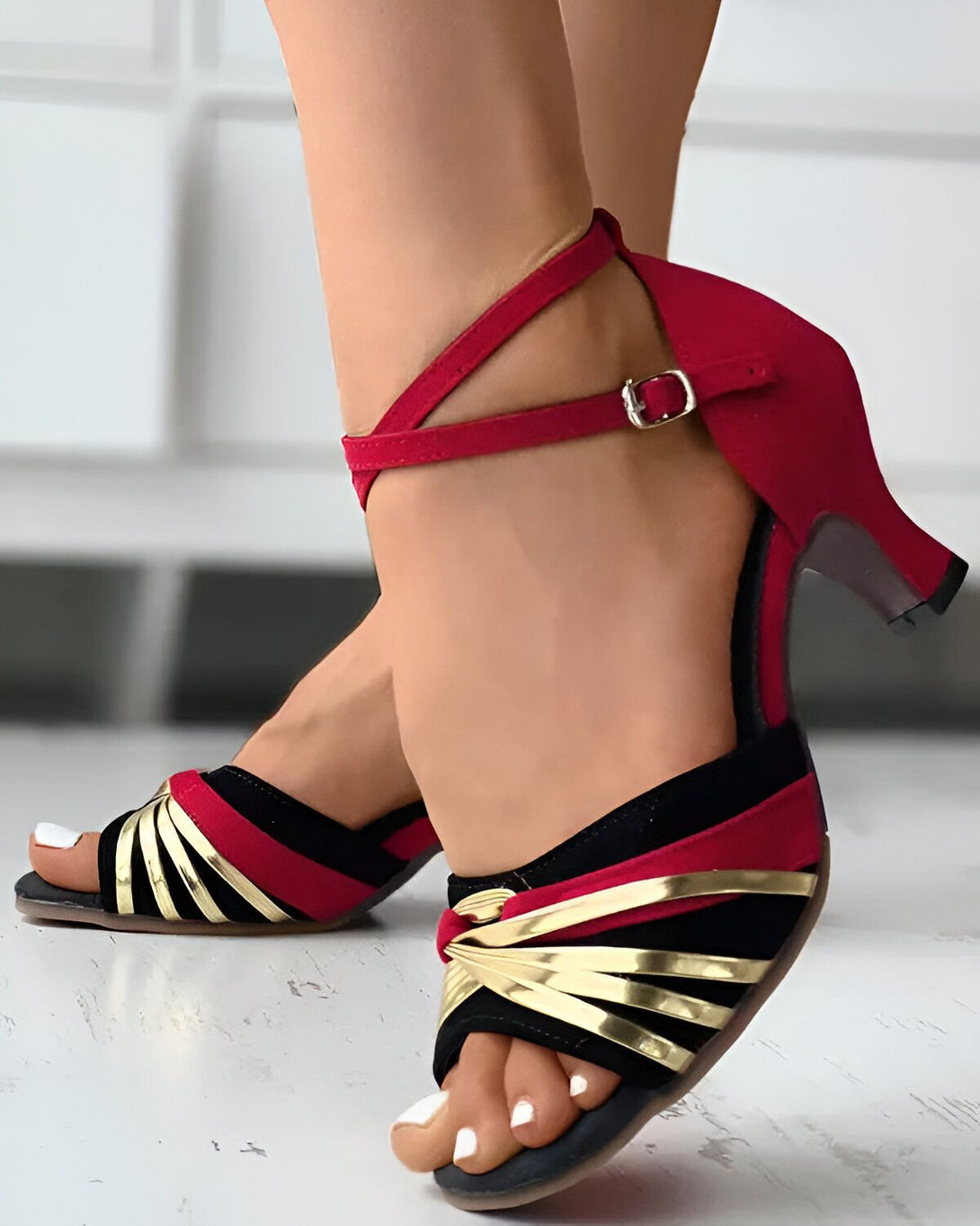 Une femme porte des sandales rouges à talons hauts. Les sandales sont ouvertes au niveau des orteils et ont une bride qui s'enroule autour de la cheville. La femme a les ongles des pieds peints en blanc.