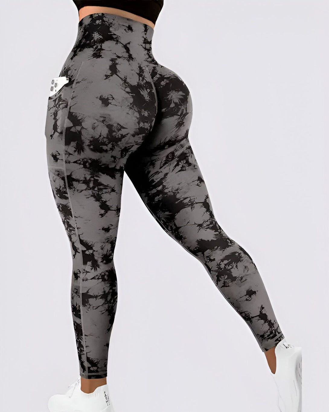 Une femme porte un legging de sport taille haute gris et noir avec une poche pour téléphone portable sur la cuisse droite. Le legging est moulant et épouse les formes du corps. Il est fabriqué dans un tissu extensible et confortable. La femme est vue de dos et porte des baskets blanches.