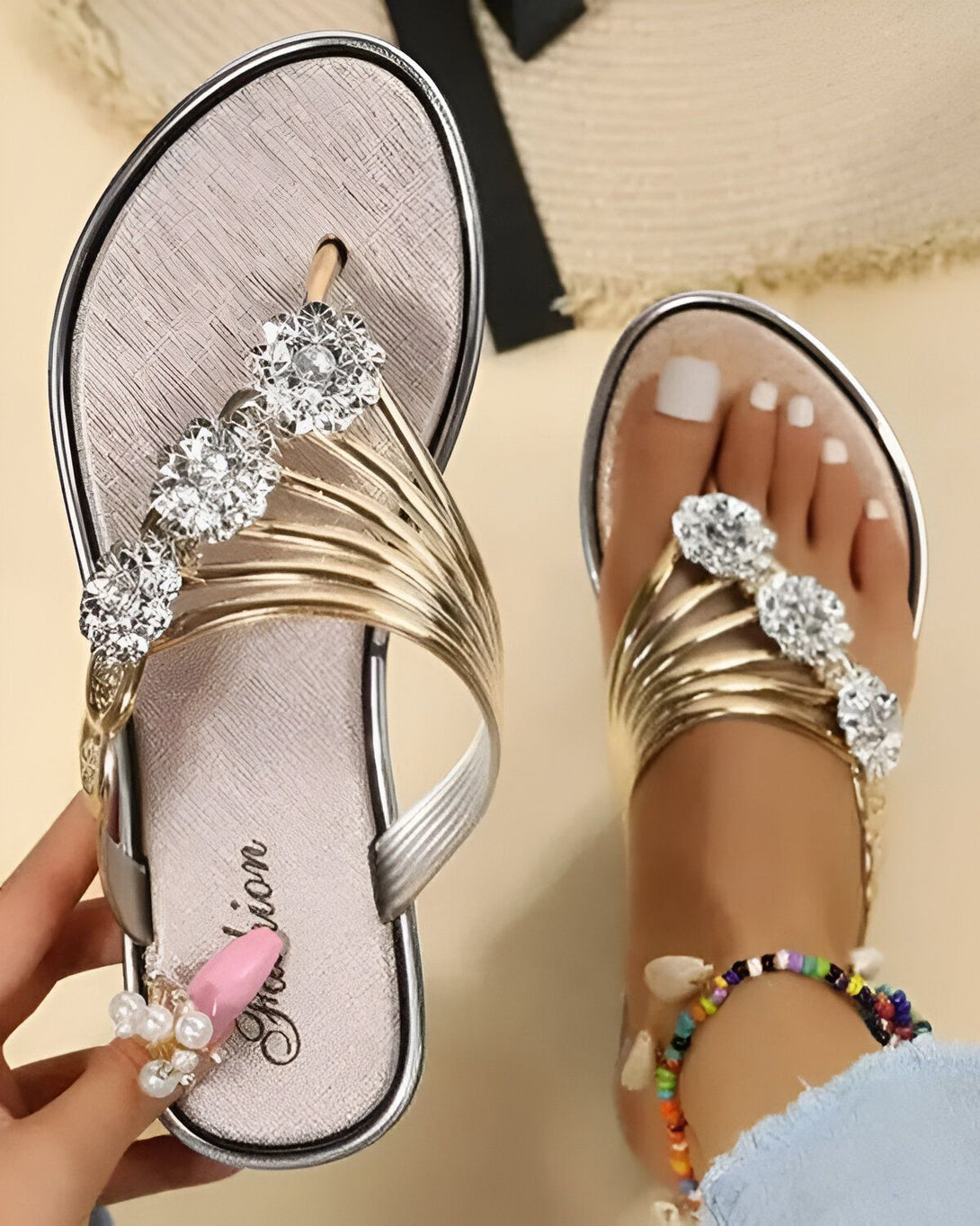 Une paire de tongs argentées avec des fleurs en strass dorées. Les tongs sont portées par une femme qui a les ongles peints en rose et porte un bracelet de cheville en perles.