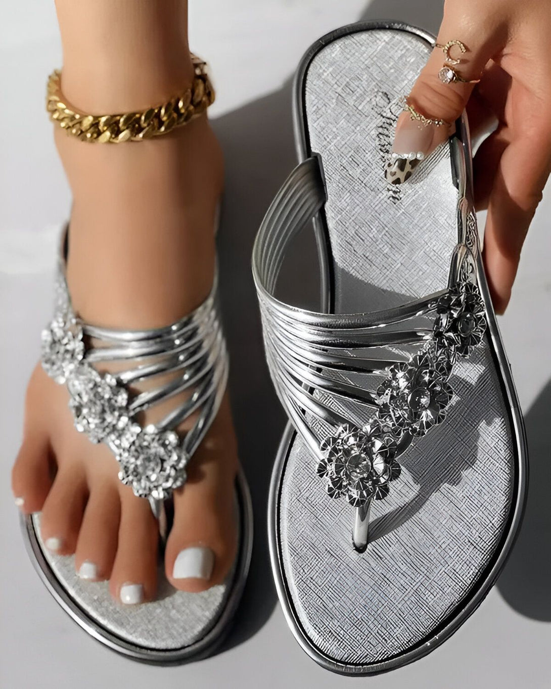 Une femme porte des tongs argentées avec des fleurs en strass. Les tongs sont faites de fines lanières de cuir argenté et les fleurs sont faites de strass blancs et argentés. La femme a les ongles des pieds peints en blanc et porte un bracelet de cheville en or.