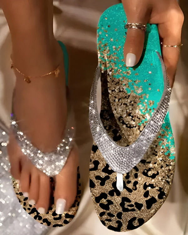 Une femme porte des tongs bleues et argentées à paillettes. Les tongs ont un motif léopard sur la semelle. La femme a les ongles des pieds peints en blanc. Elle porte également un bracelet de cheville en or.