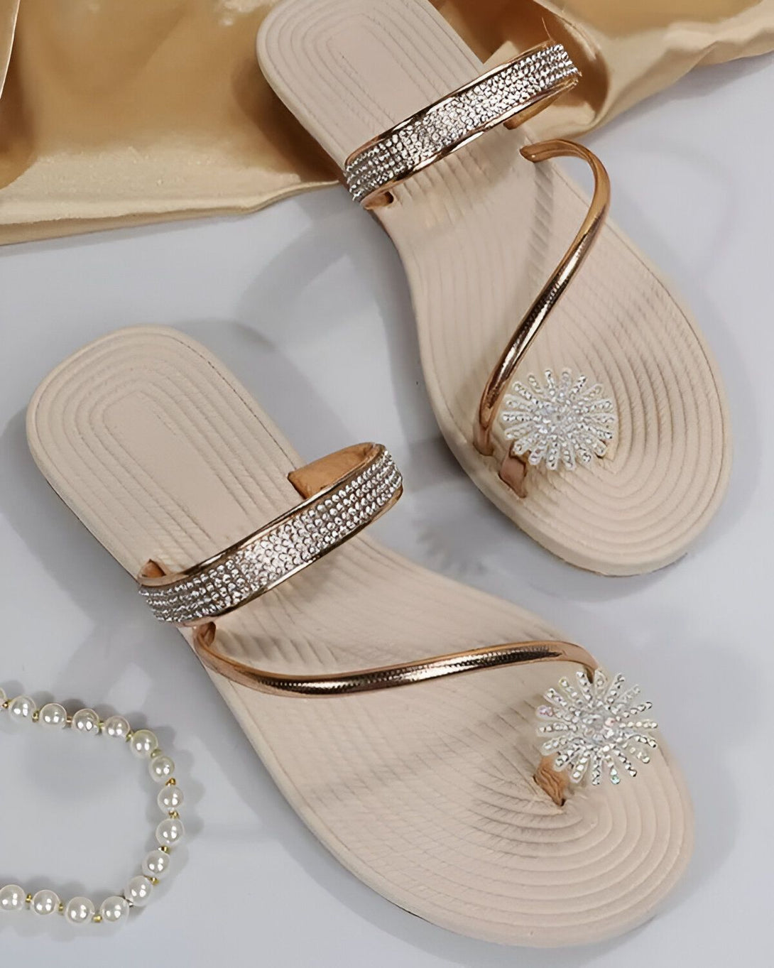 Une paire de tongs beige et doré avec des strass sur le dessus et une fleur en strass sur le bout. Les tongs sont sur un fond blanc avec un tissu doré froissé à côté.