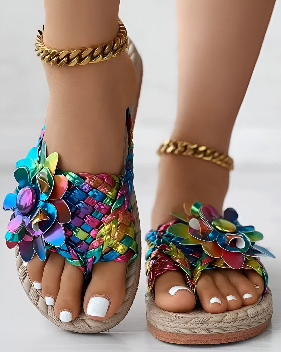 Une paire de sandales plates pour femmes avec des fleurs colorées sur le dessus. Les sandales sont de couleur marron clair et ont une semelle compensée. Les fleurs sont de couleur rose, jaune, bleu et vert. Les sandales sont portées par une femme qui a les ongles des pieds peints en blanc. La femme porte également un bracelet de cheville en or.