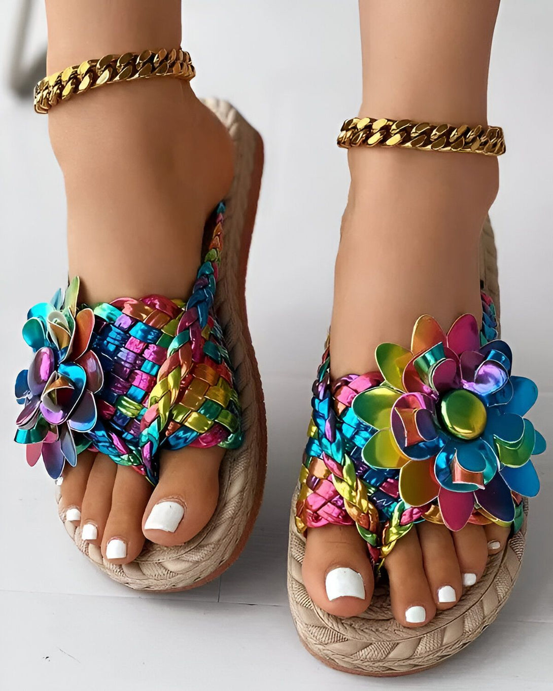 Une paire de sandales plates à enfiler pour femme avec des fleurs colorées sur le dessus. Les sandales sont de couleur beige et ont une semelle compensée. La femme porte également une chaîne de cheville dorée.