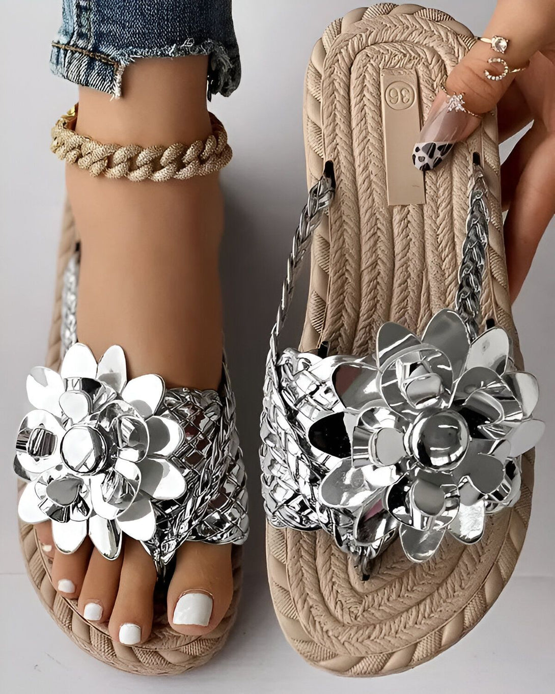 Une paire de tongs beige et argentée avec des fleurs en métal. Les tongs sont portées par une femme qui porte un jean bleu et un haut blanc. La femme est assise sur une chaise et l'on peut voir ses pieds et ses jambes.