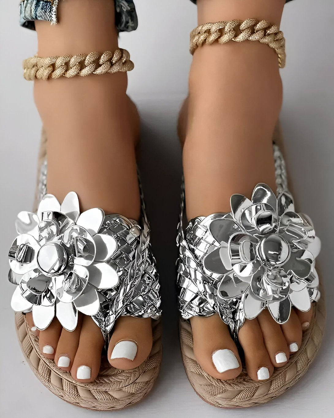Une paire de sandales plates argentées avec des fleurs en métal. Les sandales sont portées par une femme qui a les ongles des pieds peints en blanc.