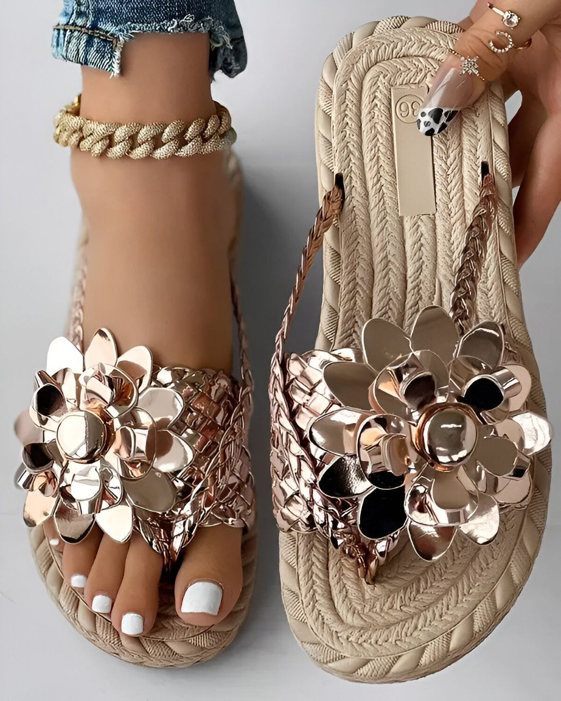 Une paire de sandales plates de couleur beige avec des fleurs en métal doré. Les sandales sont portées par une femme qui porte un jean bleu et un haut blanc. La femme est assise sur une chaise et l'on peut voir ses pieds de face.