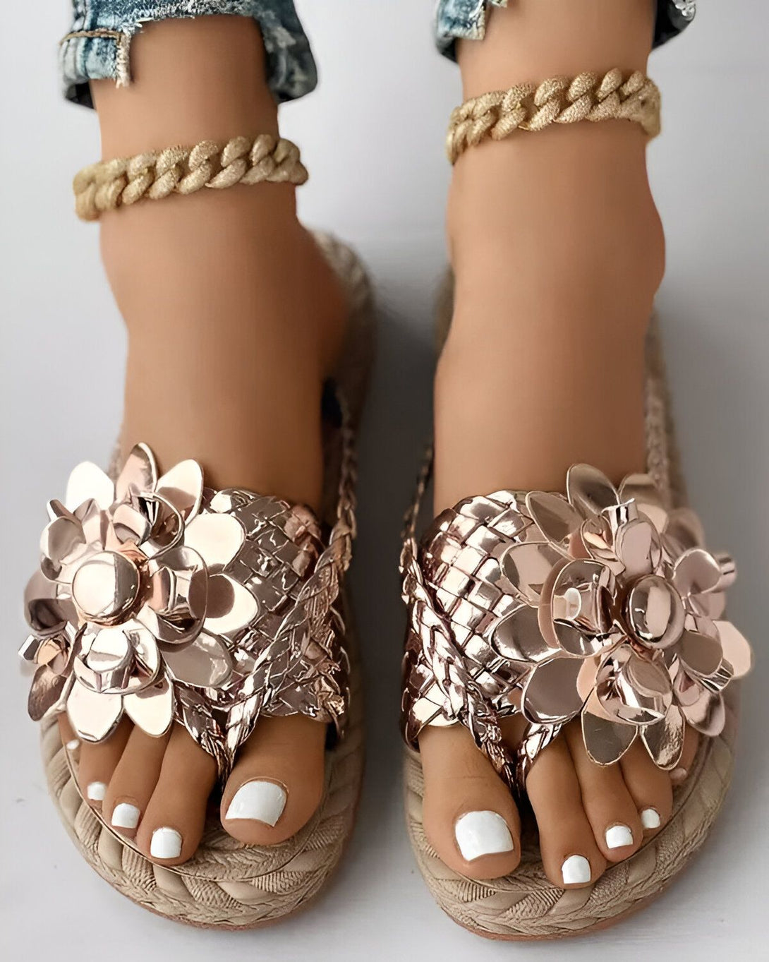 Une paire de sandales plates dorées avec des fleurs en métal. Les sandales sont portées par une femme qui porte un jean et un bracelet de cheville doré.