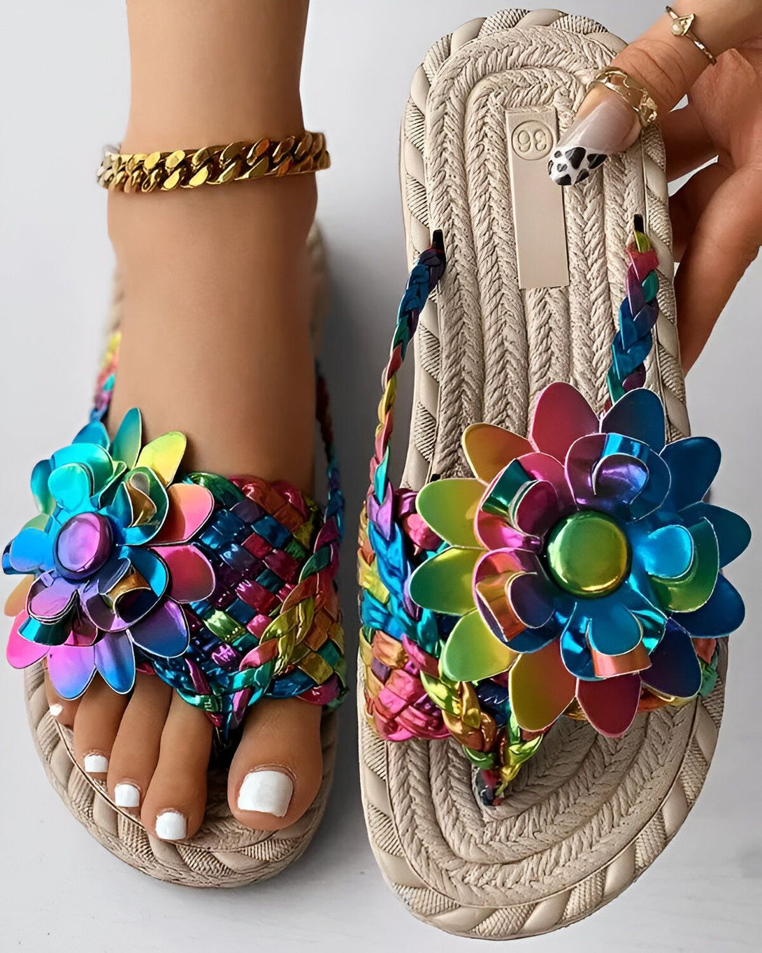 Une paire de tongs plates avec des fleurs colorées en métal. Les tongs sont de couleur beige et ont une semelle tressée. Les fleurs sont de couleur rose, jaune, bleu et vert.