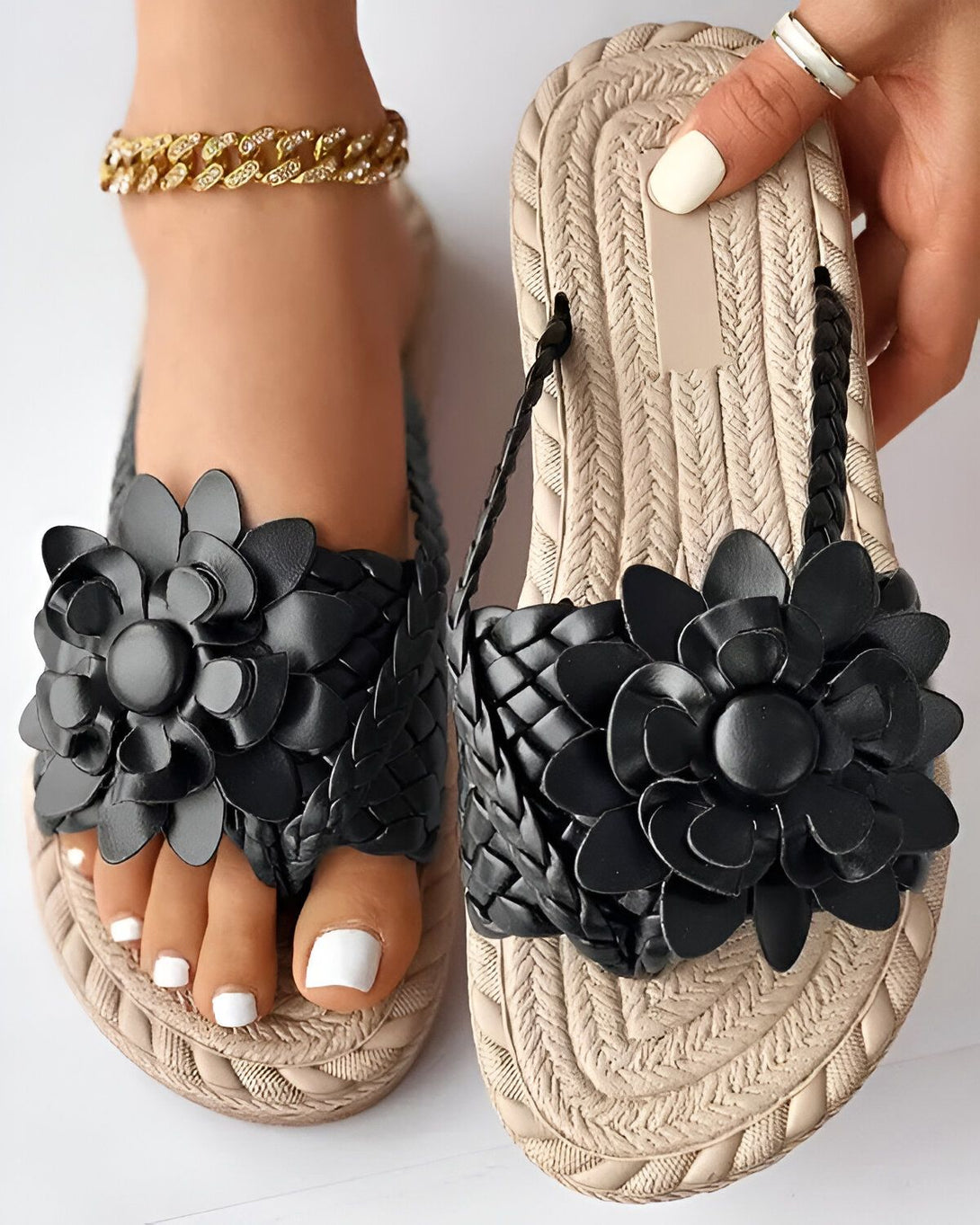 Une paire de sandales plates noires et beiges pour femmes. Les sandales sont ornées de fleurs noires en cuir. La semelle est en corde tressée beige et la bride est noire.