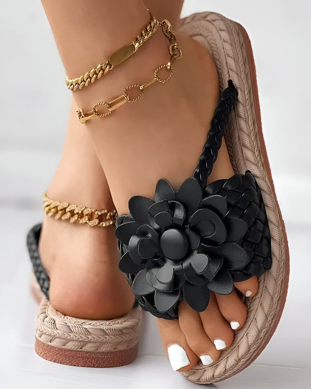 Une femme porte des sandales noires à fleurs. Les sandales sont plates et ont une bride qui passe entre les orteils. La femme a les ongles des pieds peints en blanc et porte deux bracelets de cheville en or.