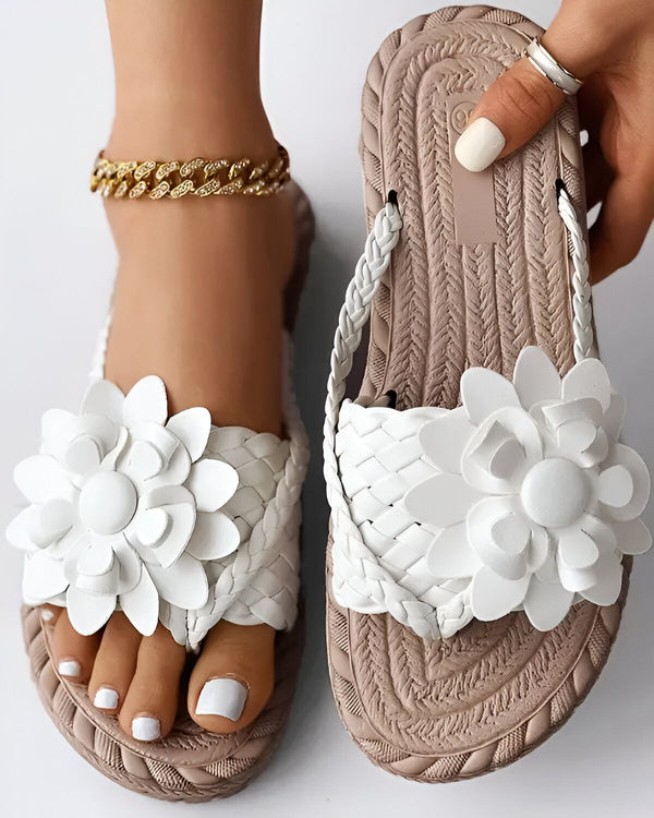 Une femme porte des sandales plates beiges avec des fleurs blanches en 3D. Les sandales sont en matière synthétique et ont une semelle tressée. La femme a les ongles des pieds peints en blanc et porte un bracelet de cheville doré.