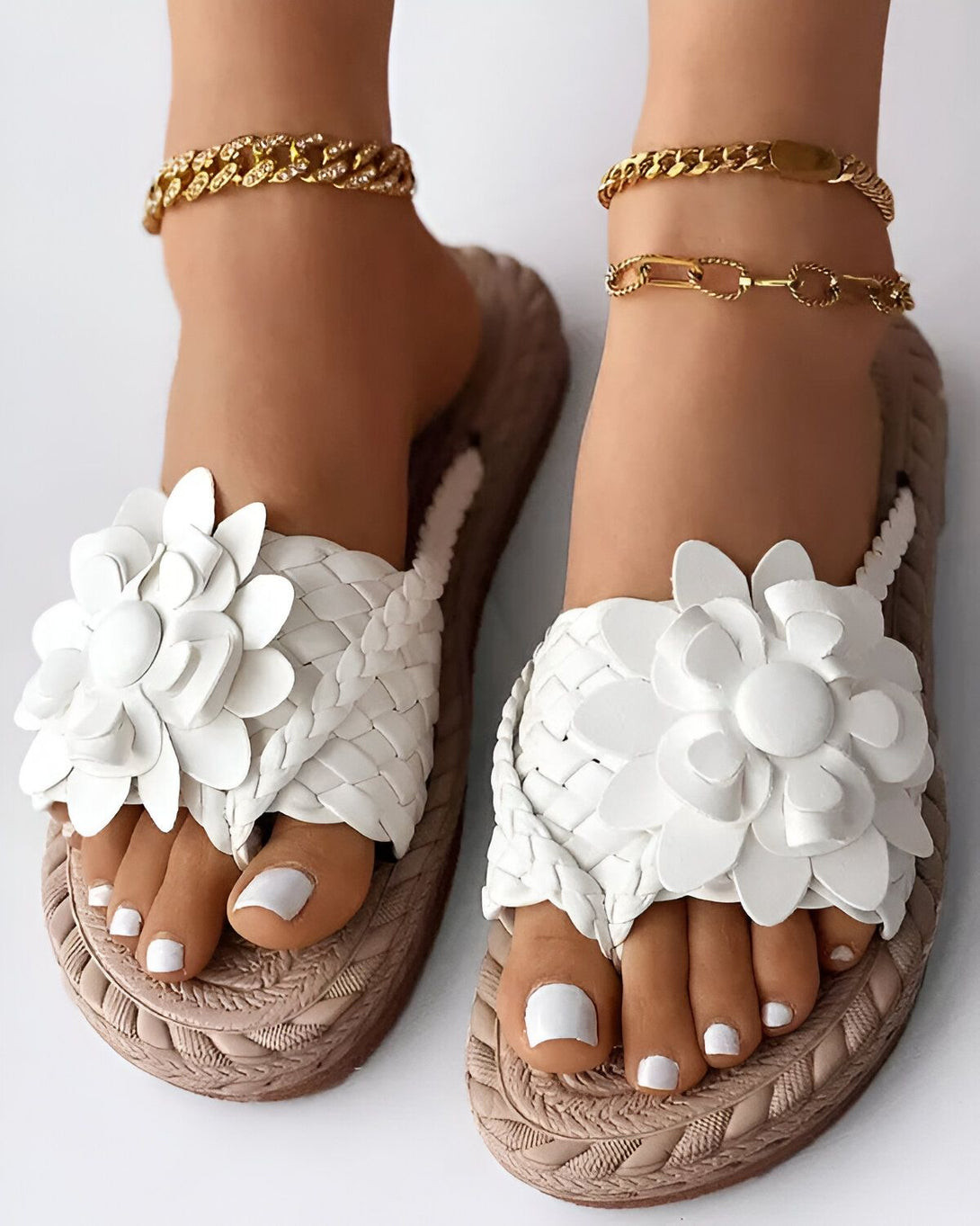 Une paire de sandales plates blanches avec des fleurs en cuir. Les sandales sont portées par une femme qui a les ongles des pieds peints en blanc et qui porte des bracelets de cheville en or.