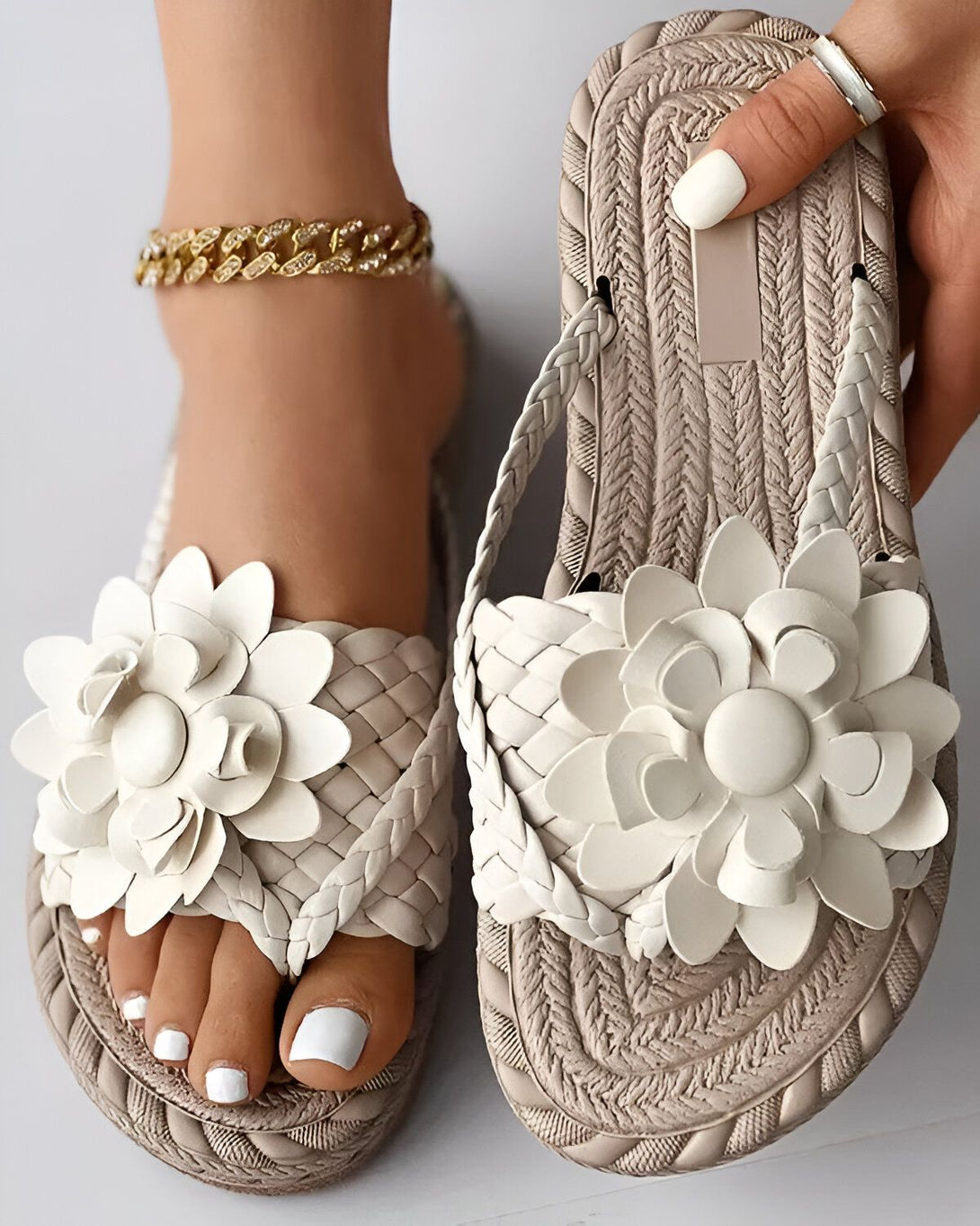 Une femme porte des sandales plates beiges avec des fleurs blanches en cuir. Les sandales sont tressées et ont une semelle épaisse. La femme a les ongles des pieds peints en blanc et porte un bracelet de cheville doré.