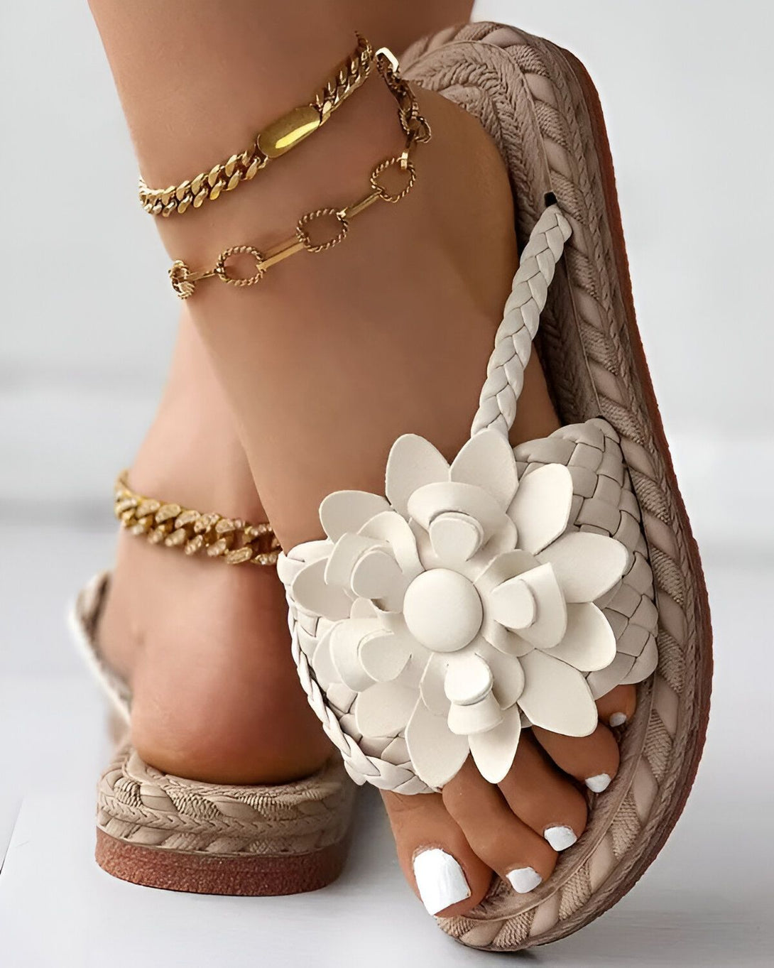 Une femme porte des sandales plates beiges avec des fleurs blanches. Les sandales sont en cuir et ont une semelle tressée. La femme porte également des bracelets de cheville en or.