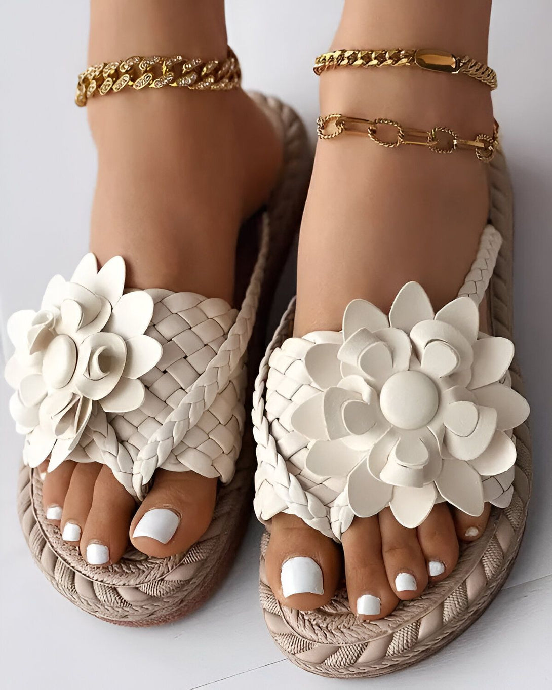 Une paire de sandales plates blanches avec des fleurs en cuir. Les sandales sont portées par un mannequin avec des chevilles fines et des ongles blancs. Le mannequin porte également des bracelets de cheville en or.