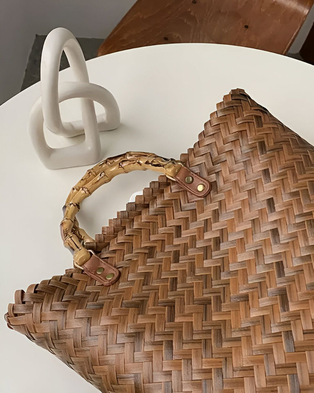 Un sac à main marron en osier avec une anse en bambou sur une table blanche à côté d'une sculpture blanche.
