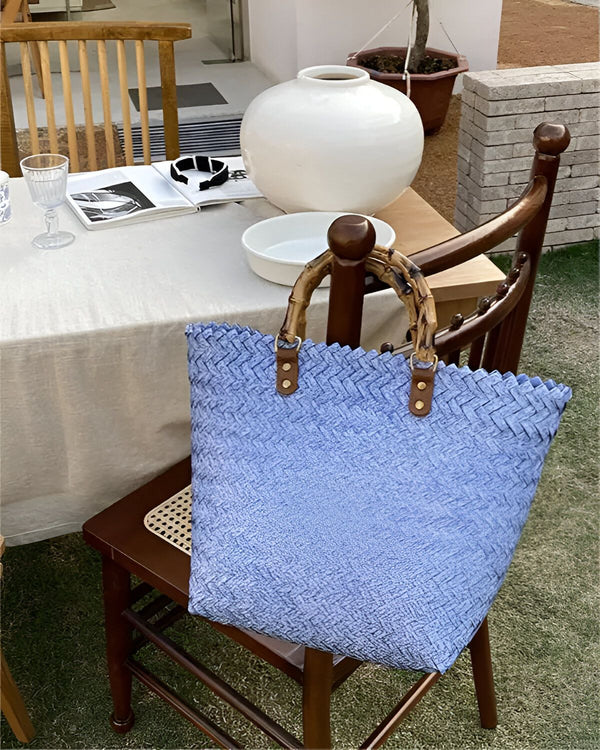 Une image d'un sac à main bleu tissé avec des anses en bambou. Le sac est posé sur une chaise en bois dans un jardin.