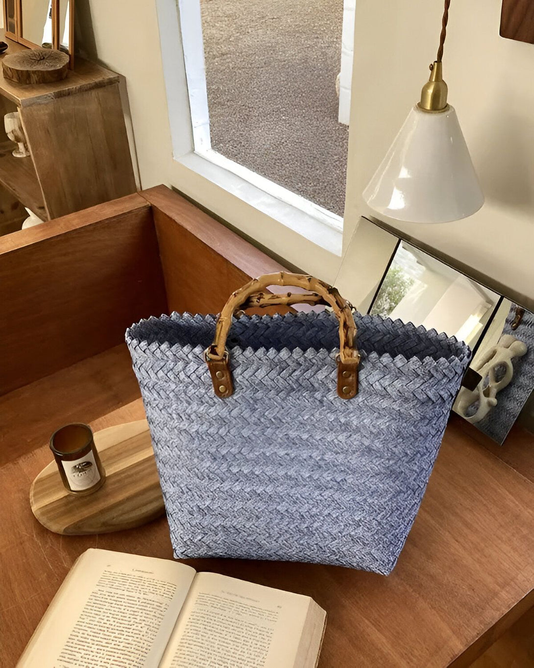 Un sac fourre-tout bleu tissé avec des anses en bambou est posé sur une table en bois. À côté du sac, il y a un livre ouvert et une bougie sur une planche à découper en bois. En arrière-plan, on peut voir une fenêtre et une lampe.