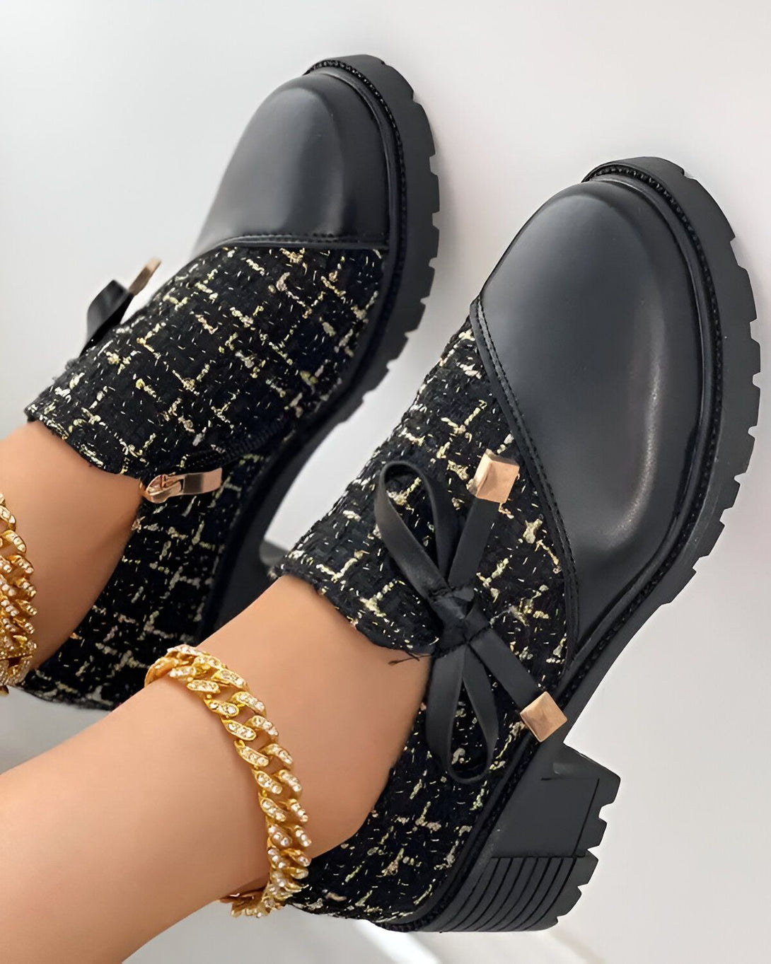 Une paire de chaussures noires à lacets avec un nœud sur le devant. Les chaussures sont portées par une femme qui a une chaîne en or à la cheville.
