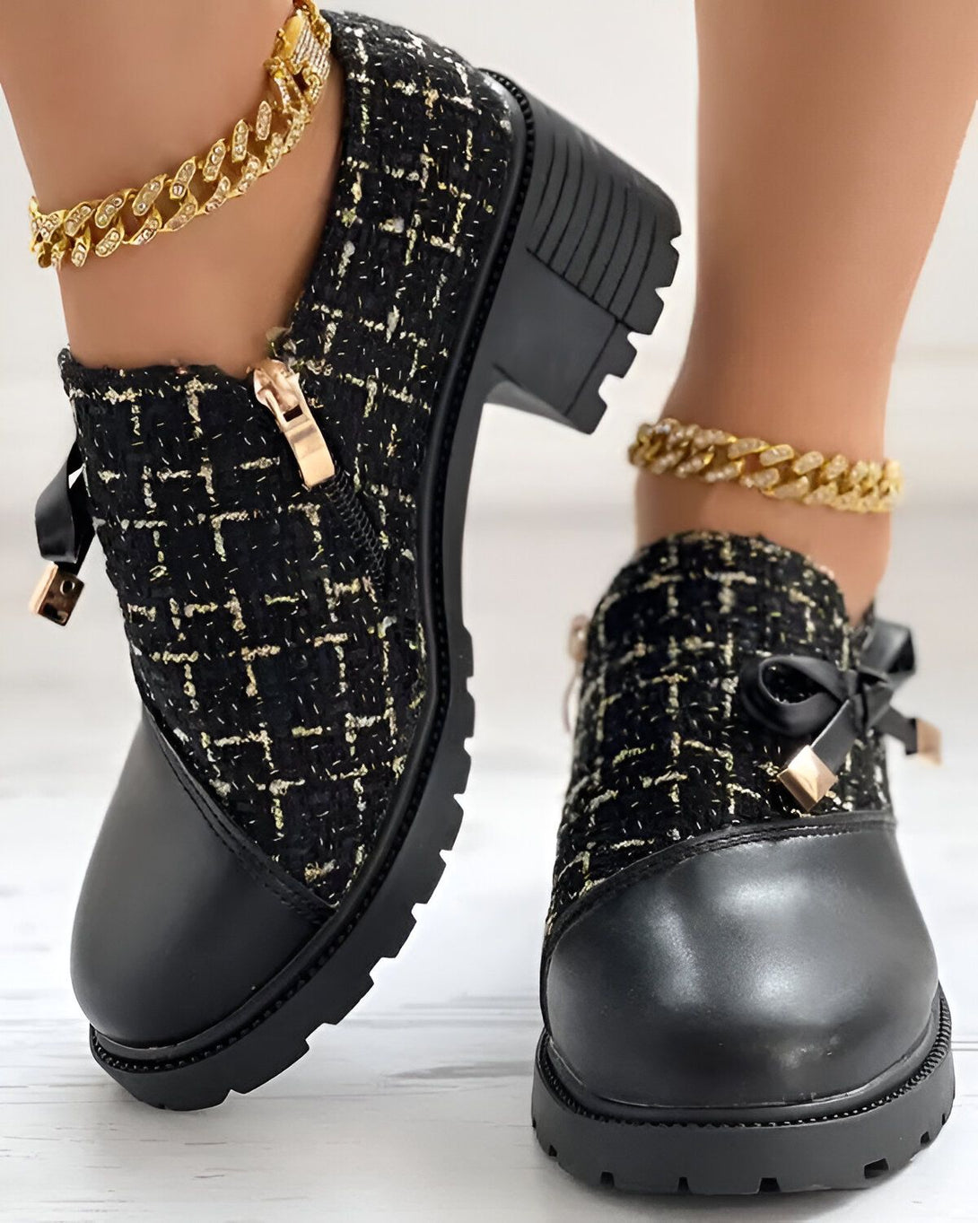 Une paire de bottines noires à talons compensés. Les bottines sont en tweed noir et ont un nœud doré sur le devant. La personne qui les porte a une chaîne dorée autour de la cheville.