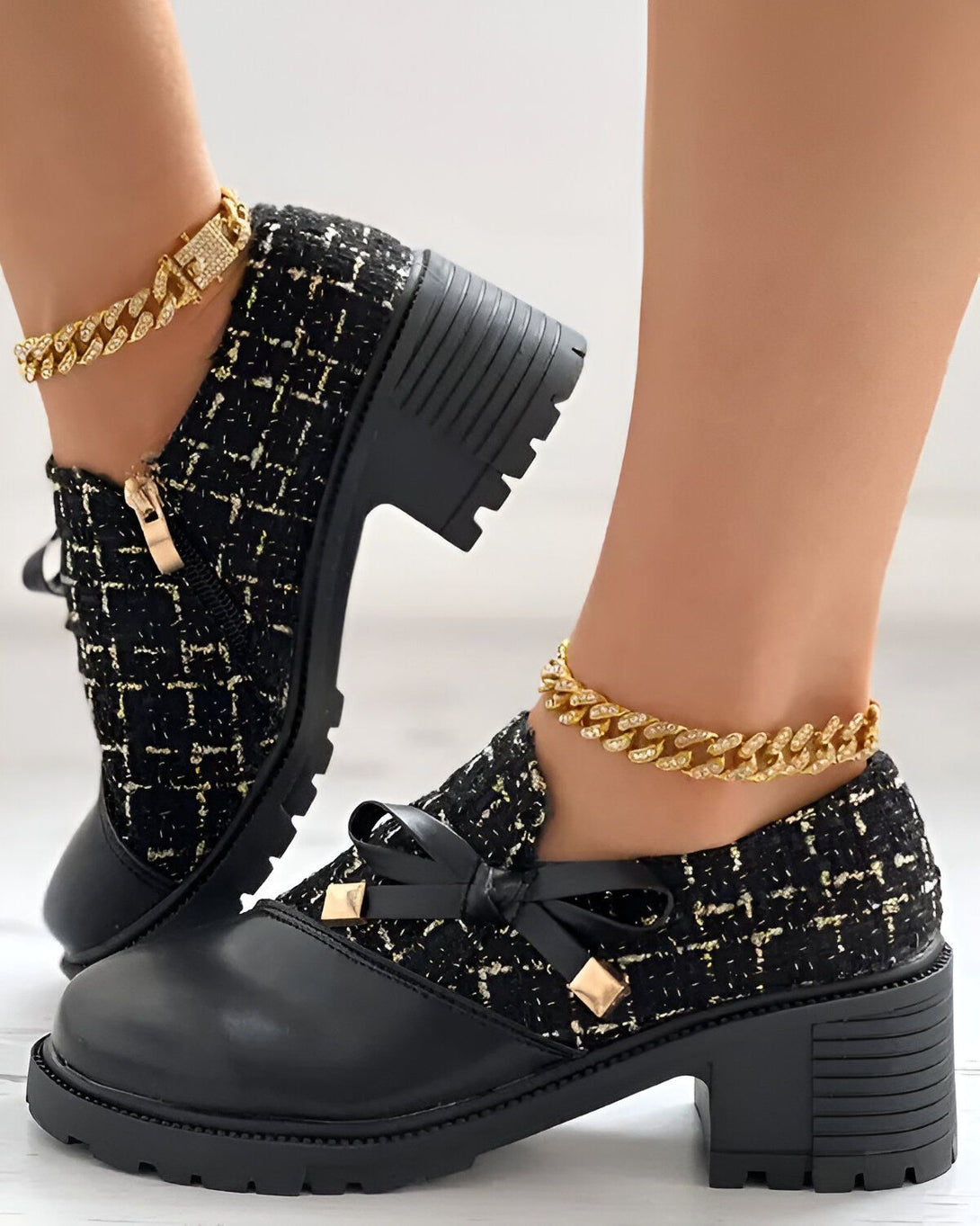 Une paire de chaussures noires à talons compensés avec des lacets et un nœud sur le devant. Les chaussures sont portées par une femme qui porte également un bracelet de cheville en or.
