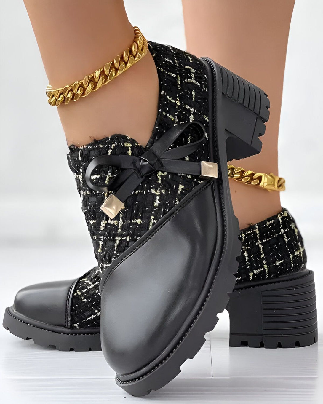 Une paire de chaussures noires à talons compensés. Les chaussures sont en tweed noir et cuir avec un nœud noir sur le devant. La personne porte également une chaîne de cheville en or.