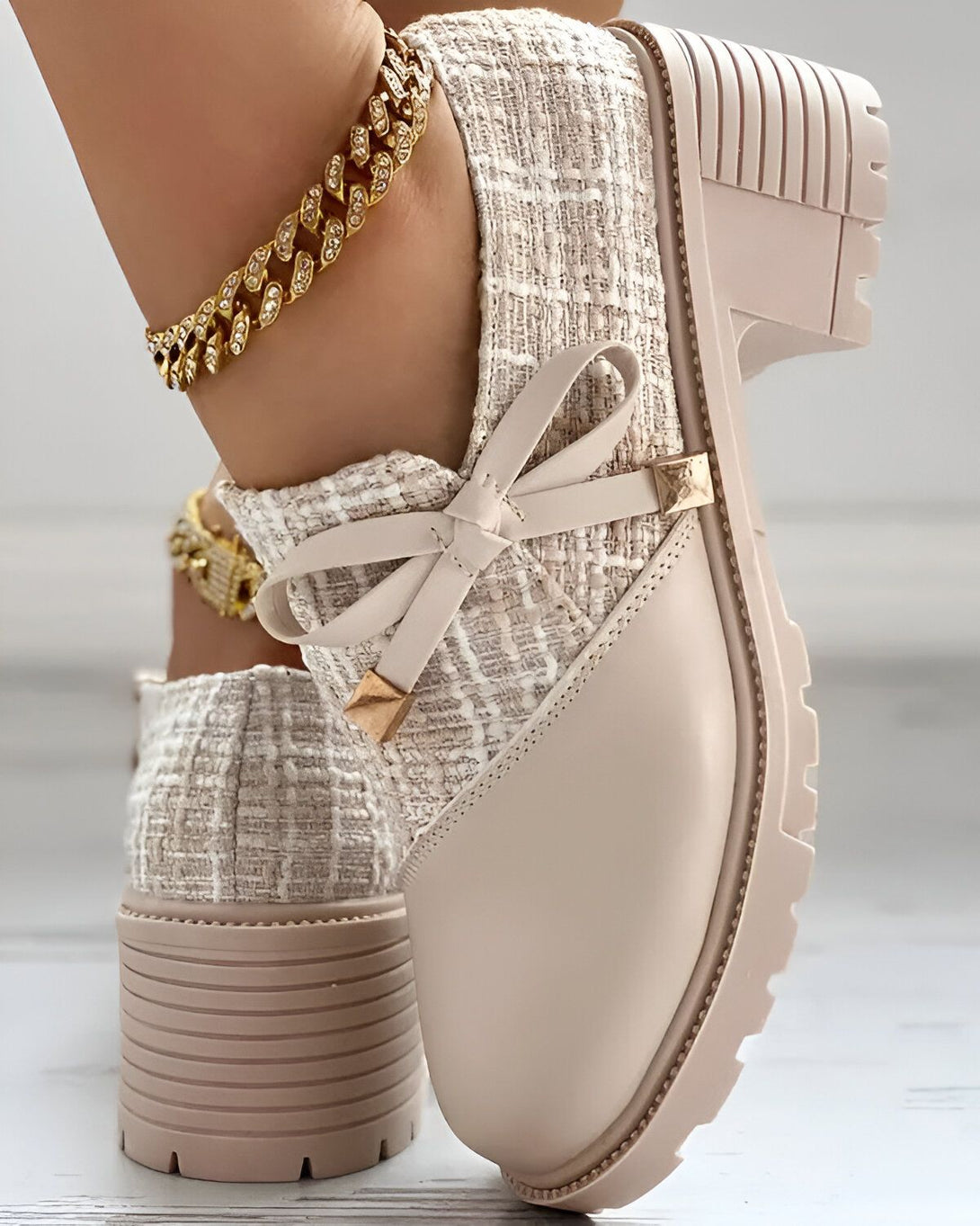 Une image d'une paire de chaussures beiges à talons compensés. Les chaussures sont faites d'un tweed beige et comportent un nœud sur le devant. Le modèle porte également un bracelet de cheville doré.