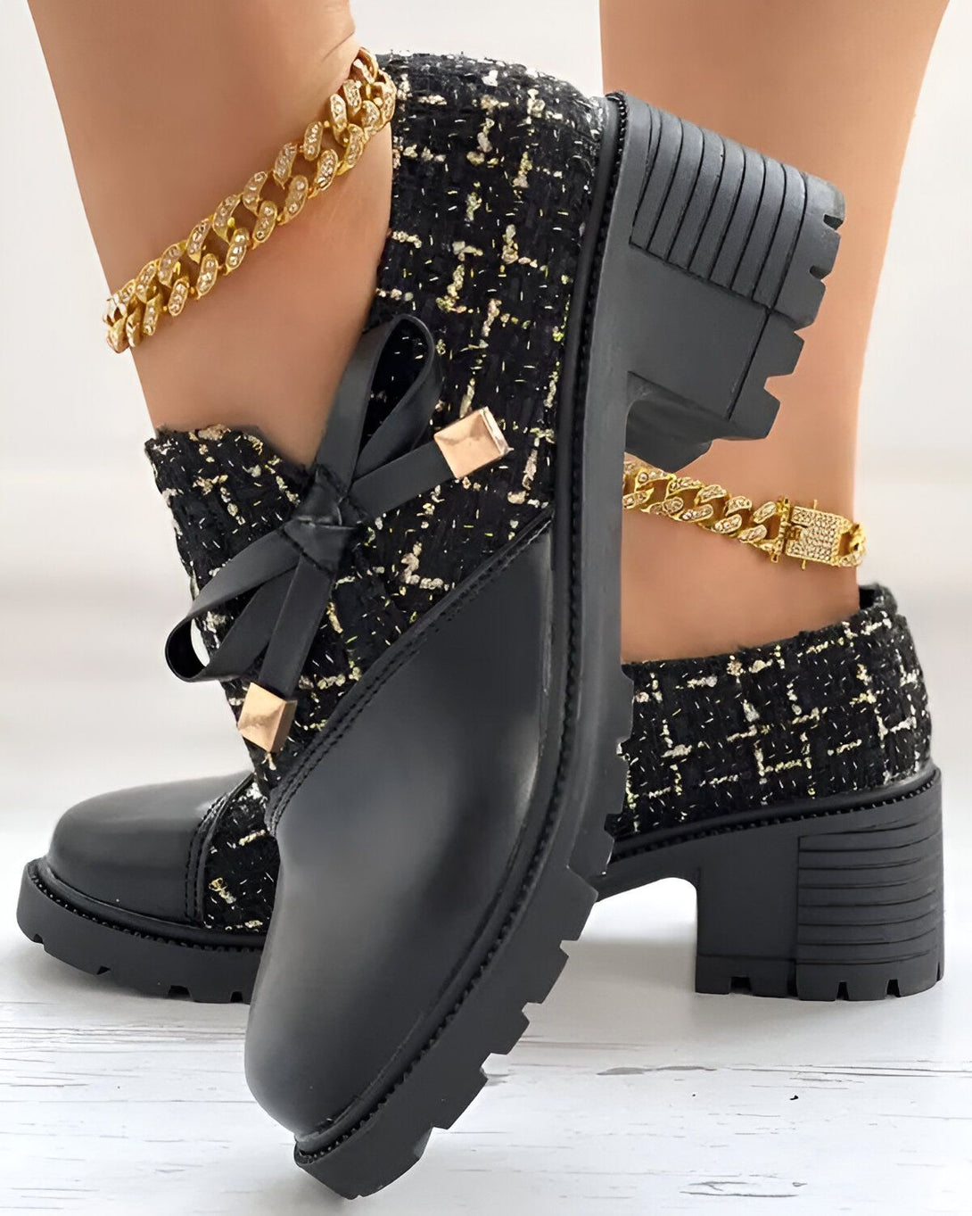 Une paire de bottines noires à talons compensés. Les bottines sont en tweed noir et cuir avec un nœud en tweed sur le devant. La personne porte également un bracelet de cheville doré.