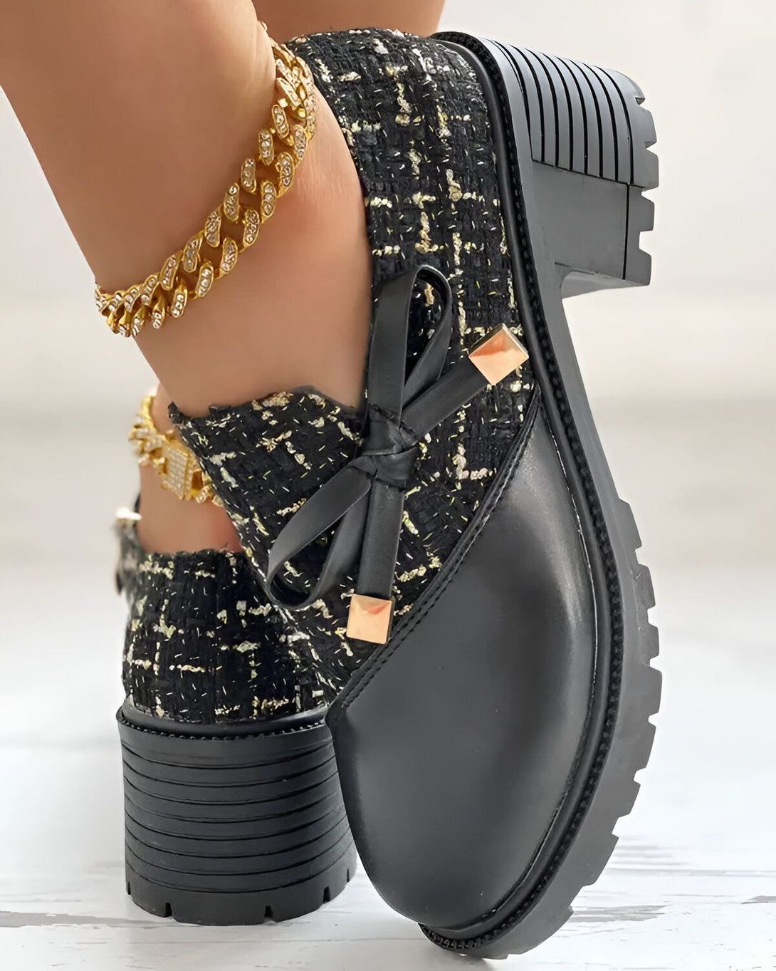 Une image d'une paire de chaussures noires à talons compensés avec un nœud en tweed noir et blanc sur le devant. Les chaussures sont portées par une femme qui porte également une chaîne de cheville en or.