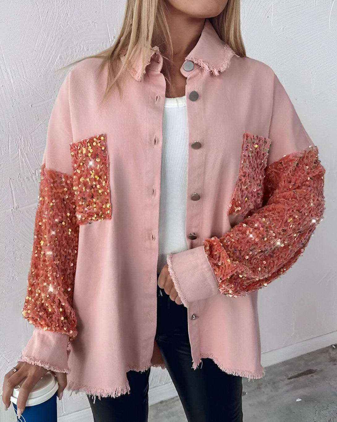 Une femme porte une veste rose en jean avec des sequins orange sur les manches et les poches. Elle porte également un pantalon noir et un haut blanc.