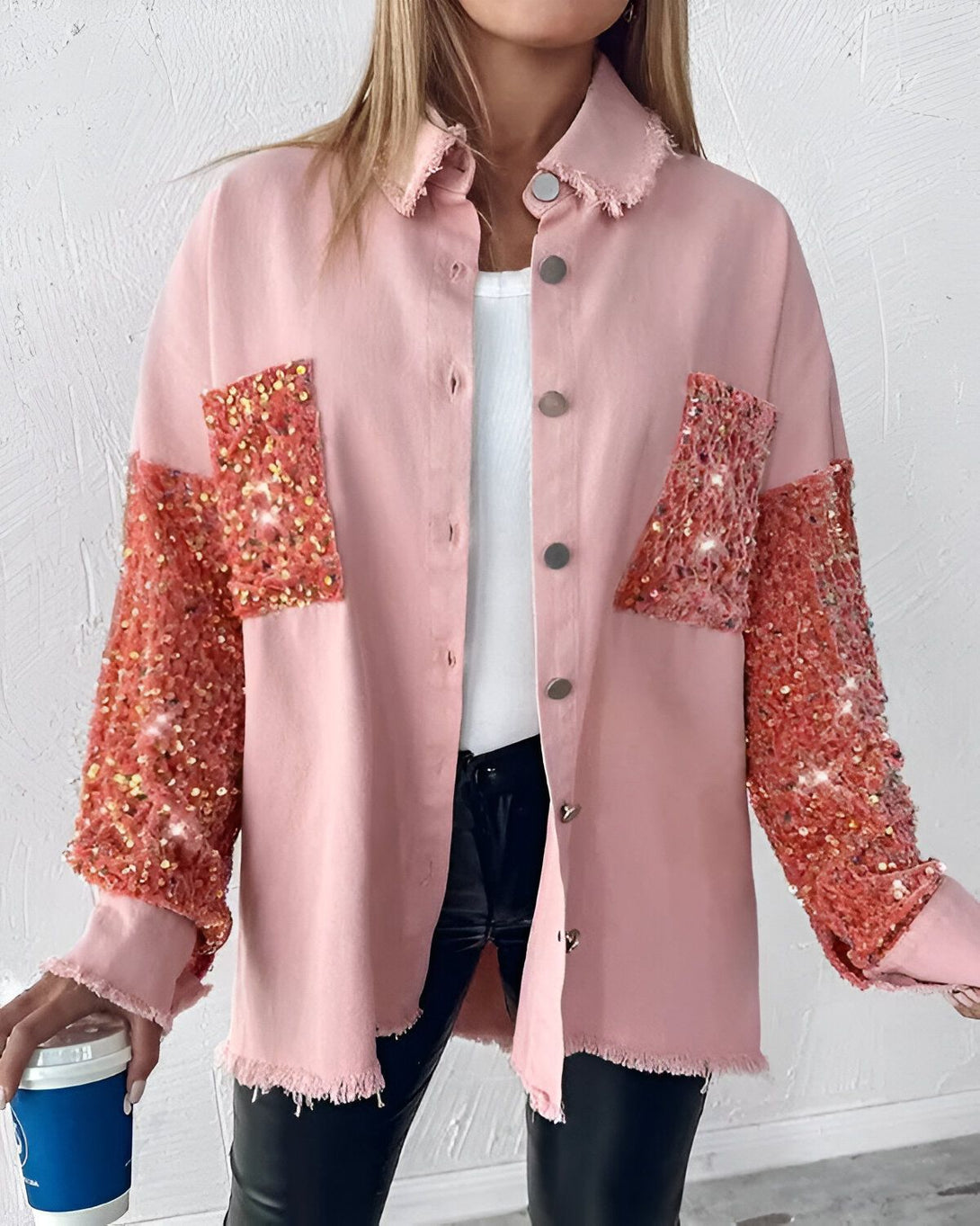 Une femme porte une chemise rose à manches longues avec des sequins orange. Elle est ouverte et on peut voir un t-shirt blanc en dessous. Elle porte également un jean noir et des talons hauts noirs. Elle a les cheveux longs et blonds et porte un maquillage léger. Elle est debout dans un café et tient une tasse à café dans sa main droite.